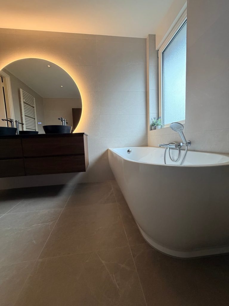 Halfvrijstaand hoekbad onder raam met donker houten wastafelmeubel, natuurstenen blad en grote ronde ledspiegel in luxe badkamer Waalre