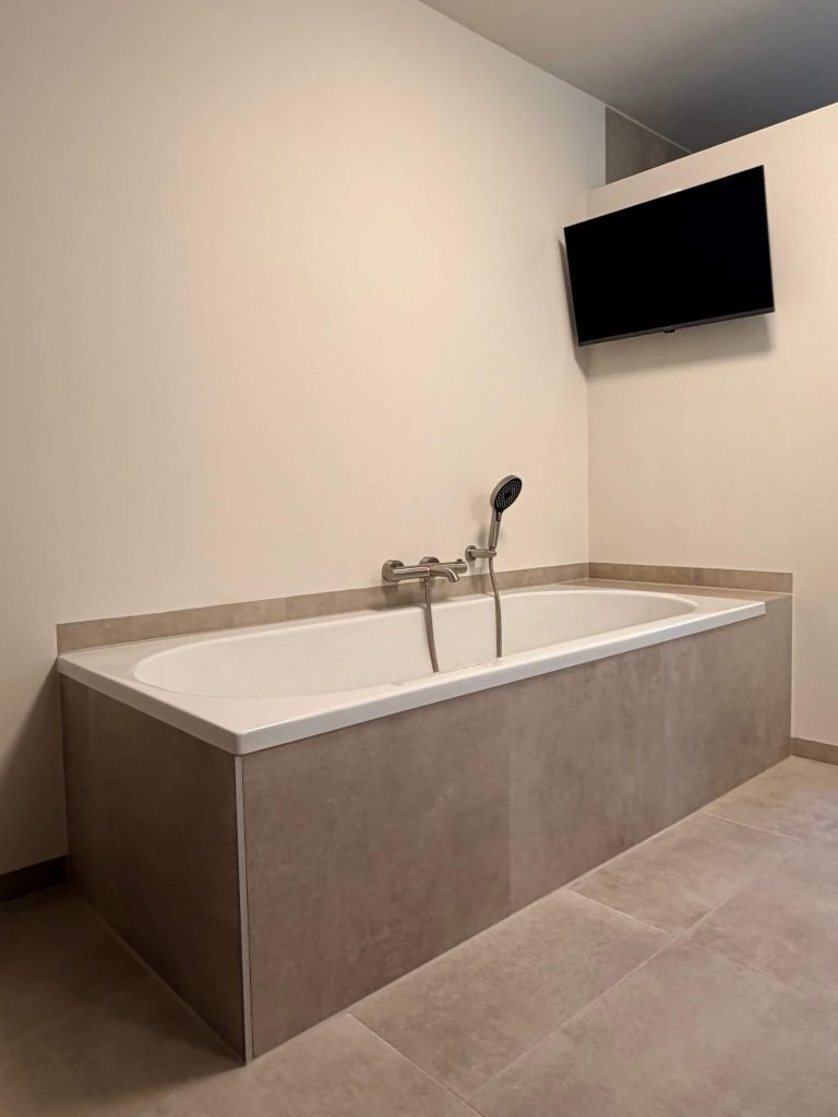 Inbouwbad omkleed met betonlook tegels in warme taupe-tint en geborsteld stalen badkraan