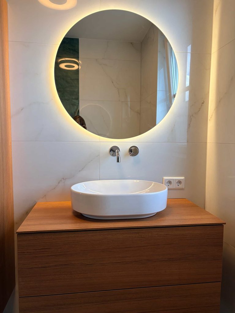 Houtlook wastafelmeubel met ronde led-spiegel en witte marmerlook in badkamer Valkenswaard